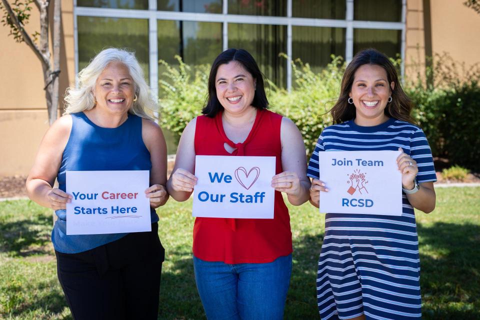 Three women standing outside, each holding a sign. One sign says "your career starts here," another sign says "We love our staff," and the last sign says "join team RCSD"