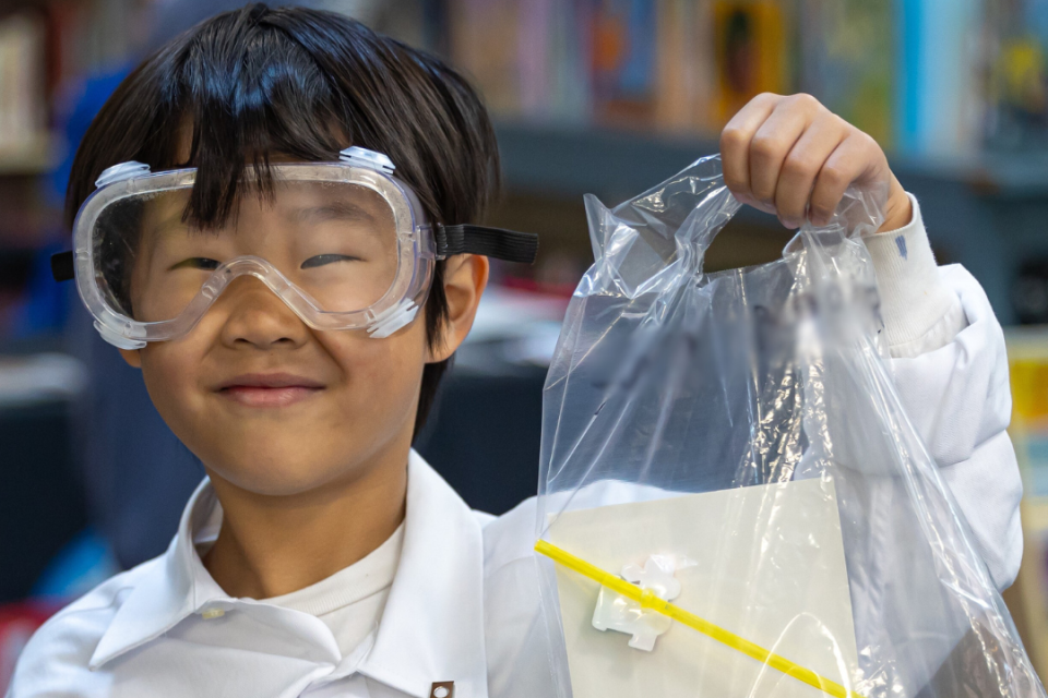 Elementary School student dressed in a lab coat and goggles stands in a library, holding up a plastic bag of school supplies.