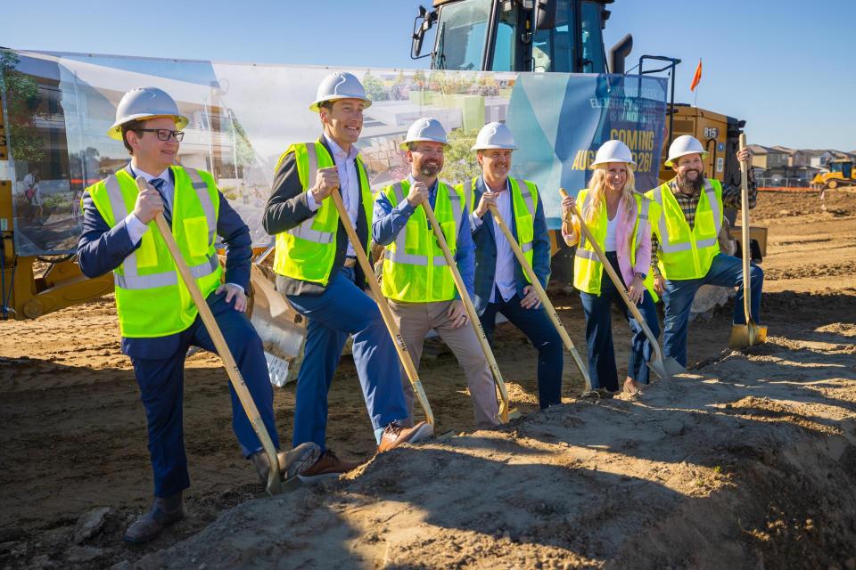 A group of people in hard hats and high-vis vests posing with shovels in front of a mound of dirt. 